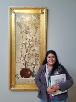 Rocio Garcia, wearing a gray jacket over a blue shirt, holds a white binder full of documents and smiles at the camera. She is standing next to a painting of pink blossoms arranged in a larger brown pot and a smaller and narrower vase. The painting has a thick gold frame with plain and ornamented decorations.